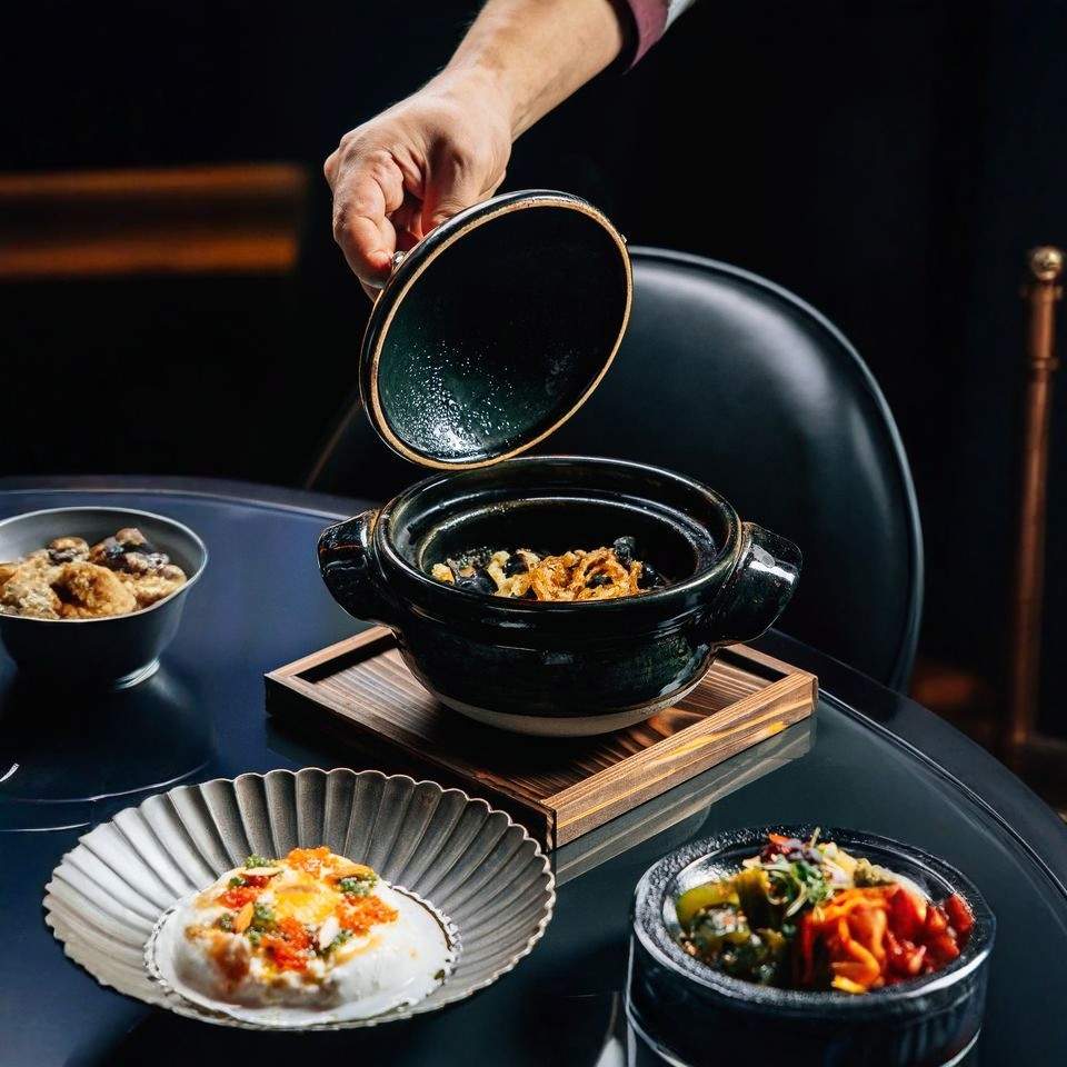 Hand opening a black clay pot with gourmet fine dining dishes served on a modern restaurant table