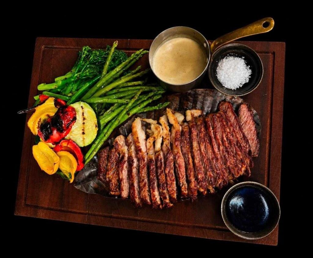 Sliced A5 Wagyu steak served with grilled asparagus, vegetables, dipping sauce, and sea salt on a wooden platter.