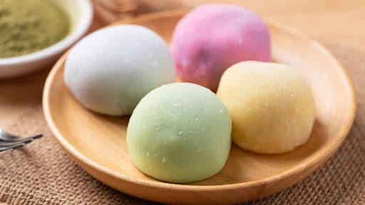 Four colorful mochi (white, pink, green, and yellow) on a wooden plate, with a bowl of matcha powder in the background.