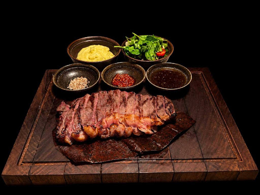 Grilled steak slices served on a wooden platter with assorted dipping sauces, hummus, and fresh greens.