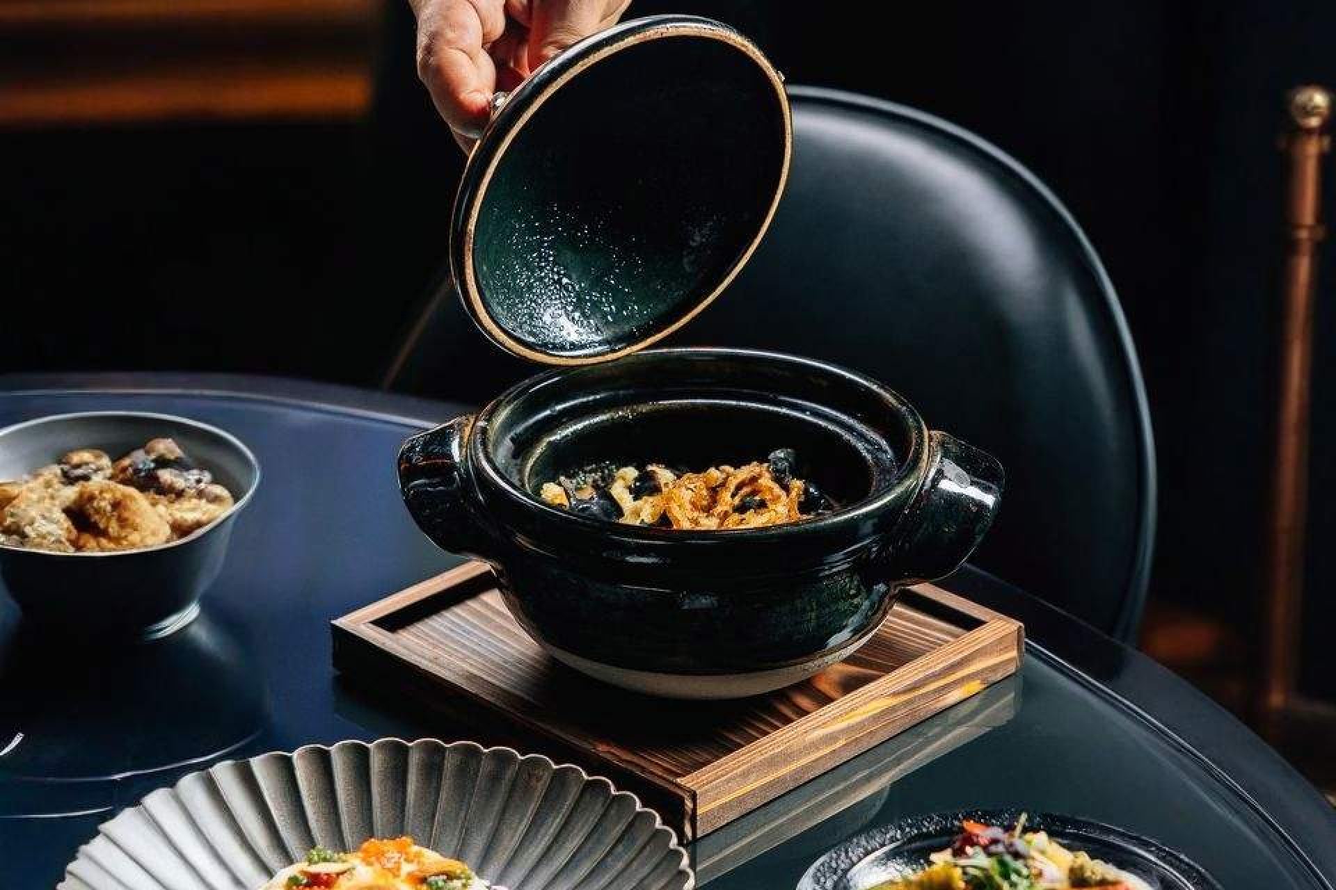 Hand opening a black clay pot with gourmet fine dining dishes served on a modern restaurant table