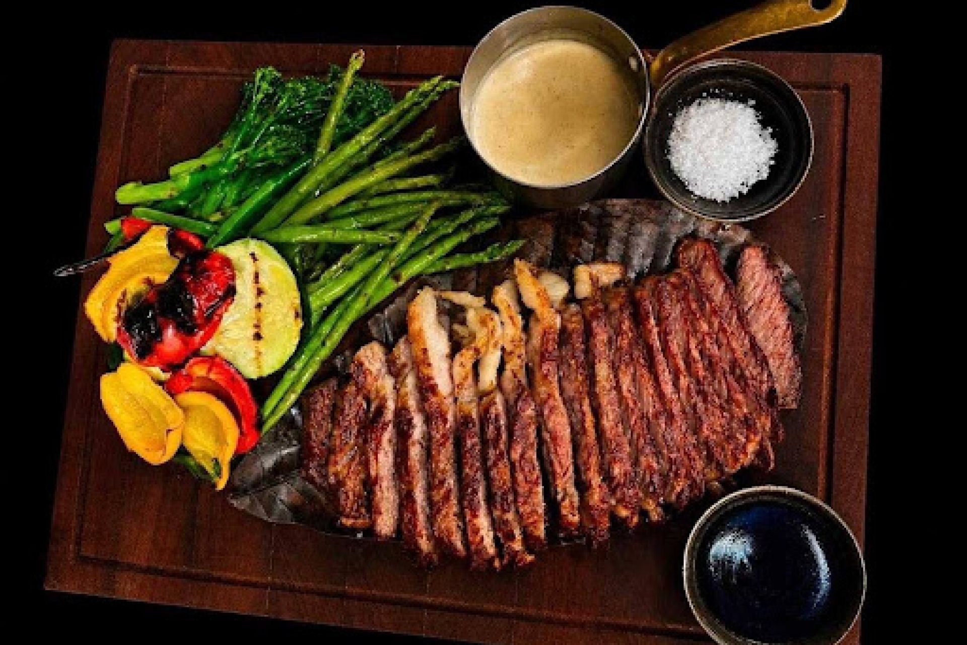 Grilled sliced steak served with green vegetables, grilled peppers, creamy sauce, and sea salt on a wooden platter.