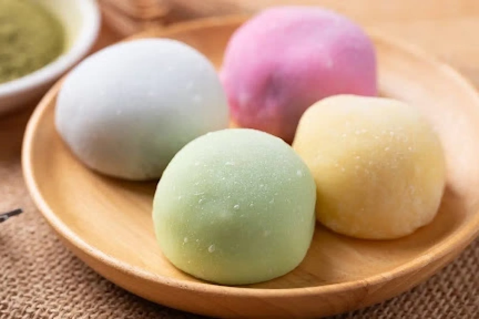 Four colorful mochi (white, pink, green, and yellow) on a wooden plate, with a bowl of matcha powder in the background.