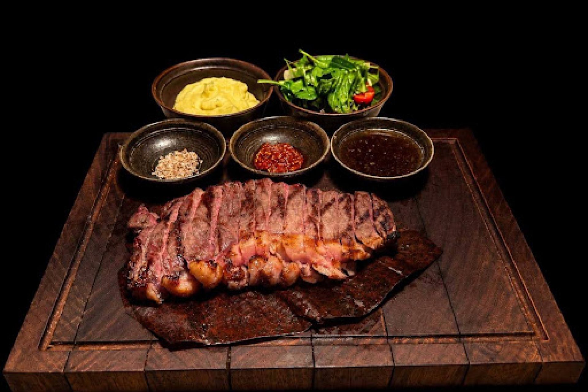 Grilled steak slices served on a wooden platter with assorted dipping sauces, hummus, and fresh greens.